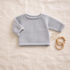 Peut inclure: Un pull pour bébé en tricot bleu clair avec des manches longues et des poignets et un ourlet côtelés. Une bague de dentition en bois et des fleurs séchées sont à côté du pull. Le pull est en fil doux.