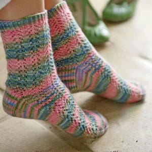 May include: Hand-knitted socks in pink, blue, and green. The socks have a patterned design and are visible from the ankles down. A pair of green shoes are in the background.