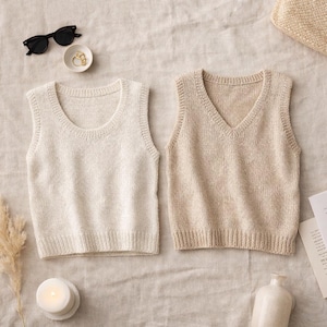 May include: Two knitted vests, one cream-coloured with a rounded neckline and the other beige with a V-neck. The vests are displayed on a neutral linen surface, alongside sunglasses, a small dish with jewellery, and a lit candle.