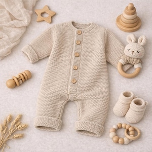 Op de afbeelding: Een beige gebreide babyromper met houten knopen, omringd door babyaccessoires. Inclusief een houten ster, een konijnenrammelaar, een houten stapelspeelgoed, een houten rammelaar en een paar gebreide babyslofjes.