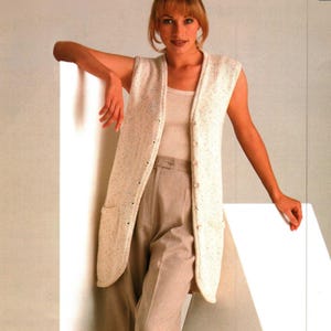 May include: A long, sleeveless, cream-colored knitted vest with a button-up front and two front pockets. The vest is worn over a light-colored top and paired with beige pants. The overall style is casual and comfortable.