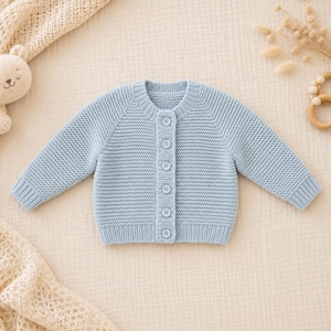 May include: A light blue long-sleeve knit cardigan featuring a round neckline and a vertical row of six matching buttons down the centre. The garment is constructed with a textured garter stitch pattern, ribbed cuffs, and a ribbed hemline.