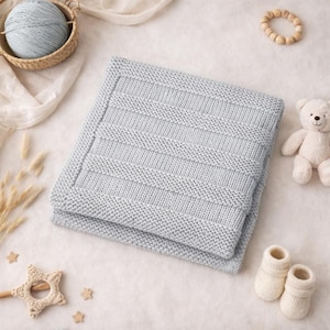 Peut inclure: Une couverture de bébé tricotée bleu clair avec un motif côtelé, pliée avec soin. Un petit panier avec de la laine bleu clair, un ours en peluche, des chaussons de bébé et un anneau de dentition en bois sont disposés autour de la couverture.
