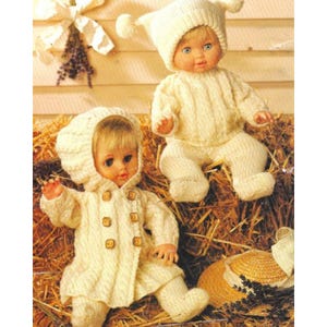 May include: Two dolls in cream-coloured knitted outfits. One doll wears a hooded coat with wooden buttons, the other a sweater and trousers with a matching hat. They are on a bed of hay, with a straw hat in the background.