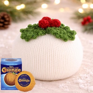 May include: A white knitted Christmas pudding, adorned with green holly and red berries. A Terry's Chocolate Orange box and a chocolate orange are placed nearby, creating a festive scene.