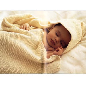 May include: A sleeping baby wrapped in a cream-colored knitted blanket with a hood. The baby's face is visible, with closed eyes and a peaceful expression. The blanket has a textured appearance, and the scene is set on a white sheet.