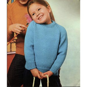 May include: A child wearing a light blue knitted jumper with ribbed cuffs and hem. Another person is partially visible in the background, wearing an orange jumper. The jumpers appear to be handmade.