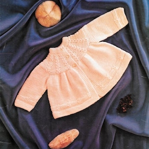 Op de afbeelding: Een lichtroze gebreid babyvestje met lange mouwen en een ronde halslijn. Het vestje heeft een decoratief patroon rond de pas en is aan de voorkant dichtgeknoopt. Het kledingstuk wordt gepresenteerd op een donkerblauwe fluwelen ondergrond.