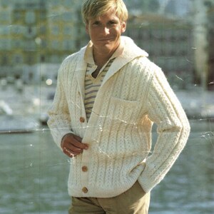 May include: A cream-coloured cable-knit cardigan with a hood and wooden buttons. The sweater has a pocket on the front and is worn over a striped shirt. The style is classic and casual.