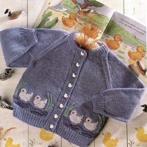 May include: A light blue knitted cardigan with a duck design. The cardigan features white duck appliques along the bottom, white buttons, and long sleeves. A children's book with duck illustrations is in the background.