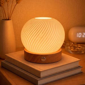 May include: A spherical, cream-colored lamp with a swirling pattern sits atop a wooden base. The lamp is placed on a stack of books on a wooden nightstand. A vase with wheat stalks is in the background.