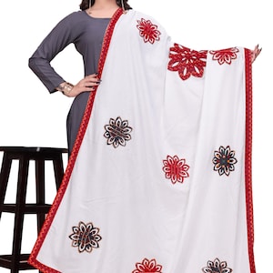 May include: A white shawl with red trim and floral embroidery. The shawl features a repeating pattern of embroidered flowers in red, black, and gold. The trim is a zig-zag pattern in red.