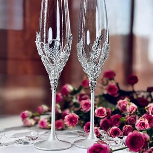 Puede incluir: Dos elegantes copas de champán con tallos plateados y intrincados adornos de cristal. Las copas de vidrio transparente presentan un diseño decorativo alrededor del cuenco. Rosas rosas y detalles de cristal sugieren un tema de boda o celebración.