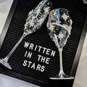 Könnte beinhalten: Zwei klare Sektgläser, verziert mit funkelnden sternförmigen Verzierungen. Die Gläser stehen auf einer schwarzen Filz-Buchstabentafel mit der Aufschrift "WRITTEN IN THE STARS". Die Gläser haben lange Stiele und stehen vor neutralem Hintergrund.