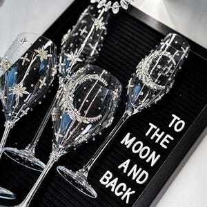 Op de afbeelding: Elegante champagneglazen met ingewikkelde kristallen versieringen. De glazen zijn versierd met ster- en bloemmotieven, en de zin "TO THE MOON AND BACK" wordt weergegeven op een zwarte achtergrond. Perfect voor een bruiloft of speciale gelegenheid.