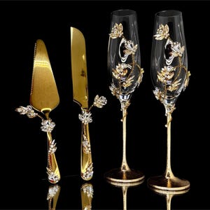 May include: A gold-colored wedding cake knife and server set with crystal and floral accents, alongside two champagne flutes. The flutes and serving set have a matching design, with gold stems and handles.