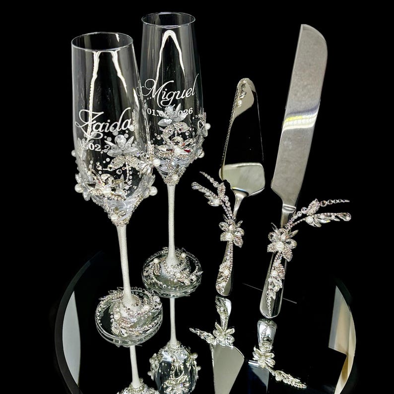 Silver jeweled wedding flutes and cake cutting set arranged on a glamorous reception head table.