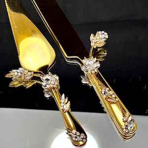 May include: A gold-colored cake server and knife set. The set features ornate floral and leaf designs with clear crystal accents. The blades are gold, and the handles are embellished with crystals and gold detailing. The set is designed for serving cake.