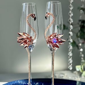 Könnte beinhalten: Zwei klare Sektgläser mit roségoldenen Flamingo-Designs. Die Flamingokörper bestehen aus glitzernden Kristallen. Die Stiele sind mit roségoldenen Strasssteinen verziert, und die Basen sind aus klarem Glas.