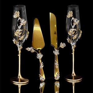 Könnte beinhalten: Ein Set aus Champagnergläsern und Torten-Servierbesteck mit goldenen und kristallenen Verzierungen. Die Gläser und das Besteck haben eine goldene Oberfläche und sind mit kristallenen Blumendesigns verziert. Das Set enthält zwei Gläser, einen Tortenheber und ein Messer.
