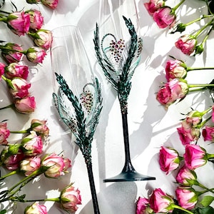 May include: Two champagne flutes with dark teal stems and bases. The clear glass flutes are decorated with green and crystal accents, forming a heart shape. Pink roses are scattered around the flutes on a white surface.