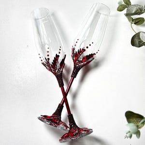 May include: Two clear glass champagne flutes featuring red crystal embellishments. The stems and bases are covered in small, dark red rhinestones, while the lower bowls are decorated with larger red leaf-shaped crystals and dots.