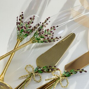 May include: Two clear glass champagne flutes with gold stems, decorated with purple and green crystal floral patterns. A matching gold cake knife and server set features similar purple and green crystal embellishments and gold ribbon bows.