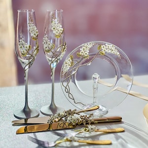 Könnte beinhalten: Zwei Champagnerflöten mit goldenen und weißen Blumenmustern, eine Kuchenteller mit einem ähnlichen Muster und ein goldenes Kuchmesser mit einem Blumenmuster.