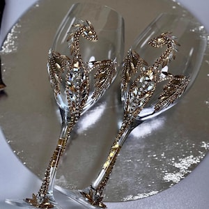 Könnte beinhalten: Zwei Weingläser aus klarem Glas, verziert mit gold- und silberfarbenen Strasssteinen in Drachenform. Die Stiele sind mit goldfarbenen Kristallen umwickelt und die Füße haben in der Mitte eine blumenförmige Kristallverzierung.
