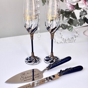 Könnte beinhalten: Zwei Sektgläser aus klarem Glas, ein Tortenmesser und ein Tortenheber mit dunkelblauen und goldenen Verzierungen. Die Gläser und Metallutensilien sind mit dem Text Mr. & Mrs. Stevenson und dem Datum 3. Januar 2027 graviert.