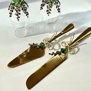 May include: A gold-colored cake server and knife set with decorative green leaves and red berry accents. The handles are adorned with gold bows and the blades are polished. Two clear champagne flutes with matching embellishments are in the background.