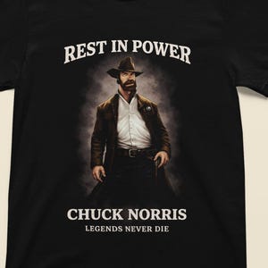 May include: Black t-shirt featuring a portrait of Chuck Norris in a hat and suit. The text "REST IN POWER" is above the image, with "CHUCK NORRIS" and "LEGENDS NEVER DIE" below. The design is centered on the shirt.