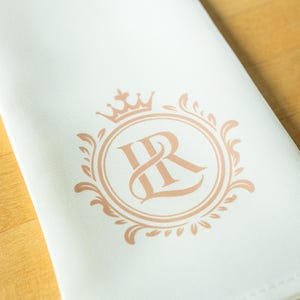 May include: Ivory-colored cloth napkin with a rose gold monogram design. The monogram features the letters "IR" within a circular frame, topped with a crown and surrounded by decorative flourishes. The napkin is folded at an angle.