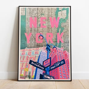 Op de afbeelding: Een ingelijste kunstdruk met een kaart van New York City en de grote roze tekst NEW YORK. Daaronder staan illustraties van rode en roze gebouwen, een blauwe lantaarnpaal en straatnaamborden voor Broadway, West 34th St en No Turns.