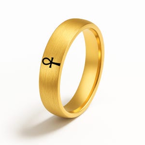 May include: A gold-colored ring with a brushed finish. The ring features a black ankh symbol on the front. The ring is a simple band design, suitable for everyday wear.