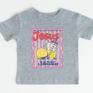 May include: A gray t-shirt featuring a colorful graphic of a glass of lemonade with a lemon slice, a straw, and the text "Jesus He Be Makin' Lemonade." The design is framed with a pink border.