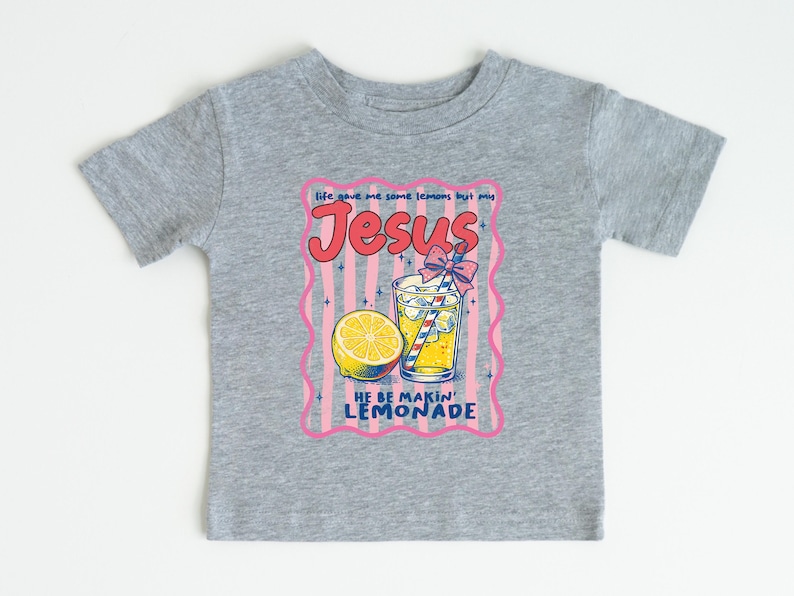 May include: A gray t-shirt featuring a colorful graphic of a glass of lemonade with a lemon slice, a straw, and the text "Jesus He Be Makin' Lemonade." The design is framed with a pink border.