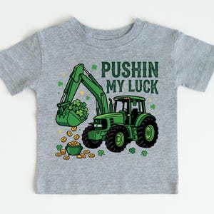 May include: A gray t-shirt featuring a green tractor with a bucket full of shamrocks and gold coins. The text "PUSHIN MY LUCK" is printed above the tractor. The design is a St. Patrick's Day theme.