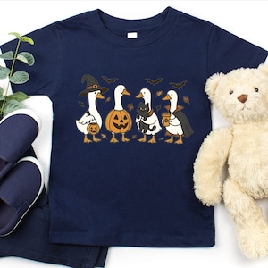 May include: Navy blue toddler t-shirt featuring a Halloween-themed graphic. The design includes four cartoon geese dressed in costumes: a witch, a jack-o'-lantern, a cat, and a vampire. The shirt is paired with navy blue pants and shoes, and a teddy bear.