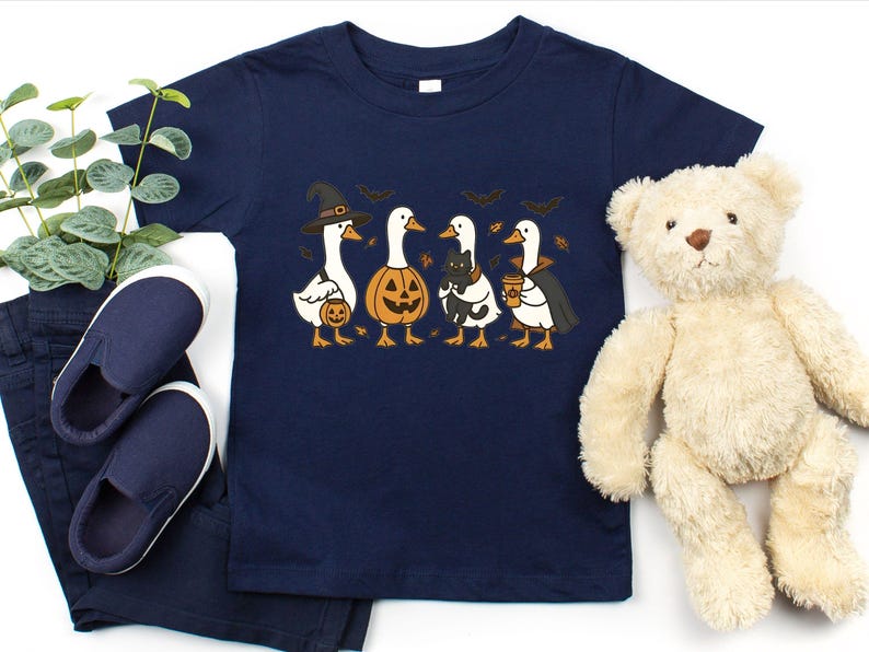 May include: Navy blue toddler t-shirt featuring a Halloween-themed graphic. The design includes four cartoon geese dressed in costumes: a witch, a jack-o'-lantern, a cat, and a vampire. The shirt is paired with navy blue pants and shoes, and a teddy bear.