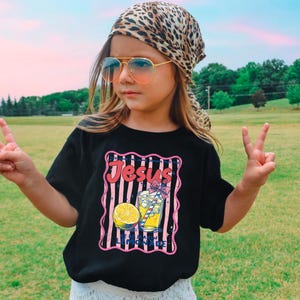 May include: A black t-shirt with a graphic that says "Jesus & Lemonade". The graphic includes a lemon, a glass of lemonade, and pink and white stripes. The person is wearing a leopard print scarf and sunglasses.