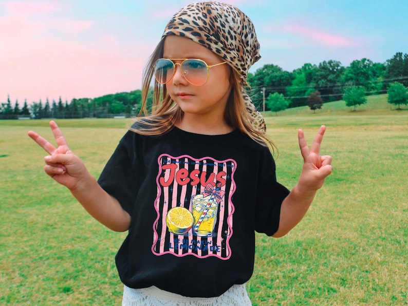 May include: A black t-shirt with a graphic that says "Jesus & Lemonade". The graphic includes a lemon, a glass of lemonade, and pink and white stripes. The person is wearing a leopard print scarf and sunglasses.