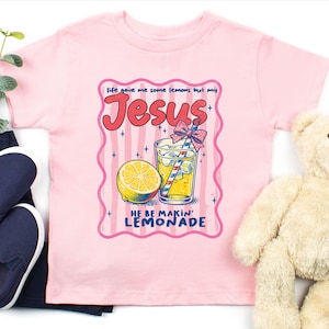 May include: Pink t-shirt with a graphic featuring a lemon, a glass of lemonade, and the text "Life gave me some lemons but my Jesus HE BE MAKIN' LEMONADE". Navy blue slip-on shoes and a teddy bear are also in the image.