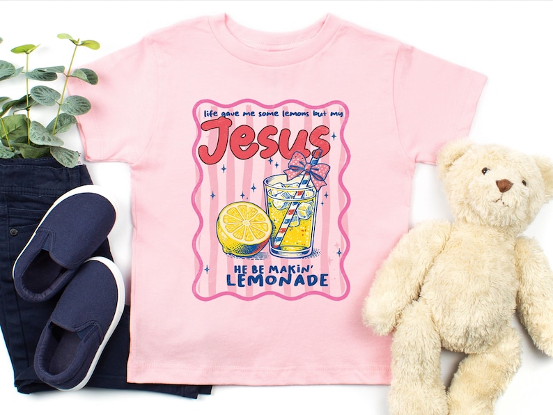 May include: Pink t-shirt with a graphic featuring a lemon, a glass of lemonade, and the text "Life gave me some lemons but my Jesus HE BE MAKIN' LEMONADE". Navy blue slip-on shoes and a teddy bear are also in the image.