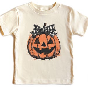 May include: Cream-colored t-shirt featuring a distressed orange pumpkin design with a black and white checkered bow. The pumpkin has a carved face with triangular eyes and a smiling mouth. A festive Halloween-themed tee.