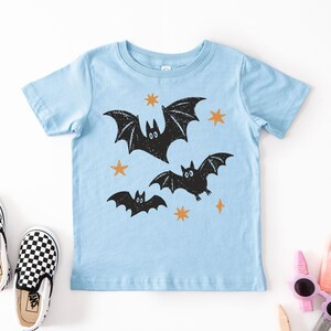 May include: A light blue short-sleeve t-shirt featuring a graphic print of three black bats in flight surrounded by small orange stars. The design has a textured, hand-stamped appearance with the bats positioned centrally on the front of the shirt.