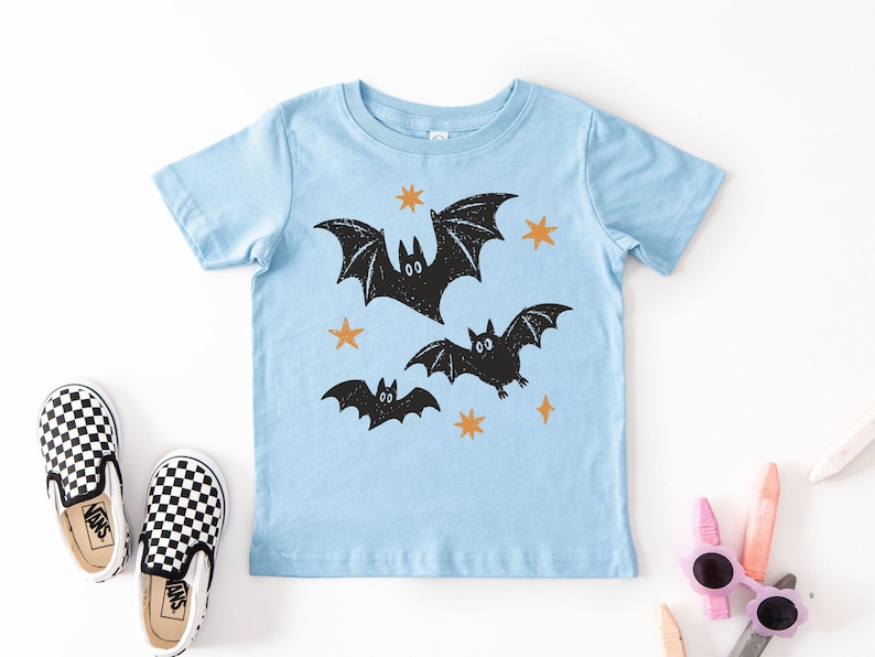 May include: A light blue short-sleeve t-shirt featuring a graphic print of three black bats in flight surrounded by small orange stars. The design has a textured, hand-stamped appearance with the bats positioned centrally on the front of the shirt.