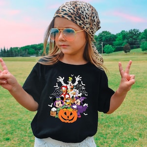 Op de afbeelding: Zwart T-shirt met een Halloween-thema met Disney-personages, waaronder Mickey en Minnie Mouse, Donald Duck en Goofy, rond een uitgesneden pompoen. Het shirt wordt gedragen door een persoon met een luipaardprint sjaal en zonnebril.