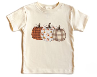 May include: Cream-colored t-shirt featuring three embroidered pumpkins. The pumpkins have different patterns: plaid, floral, and checkered. Each pumpkin has a brown stem and a twine bow. A fall-themed graphic tee.