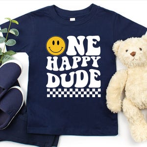 May include: Navy blue t-shirt with the text "ONE HAPPY DUDE" in white, featuring a yellow smiley face. The shirt is paired with navy blue pants and shoes, and a teddy bear. A checkered pattern is at the bottom of the shirt.